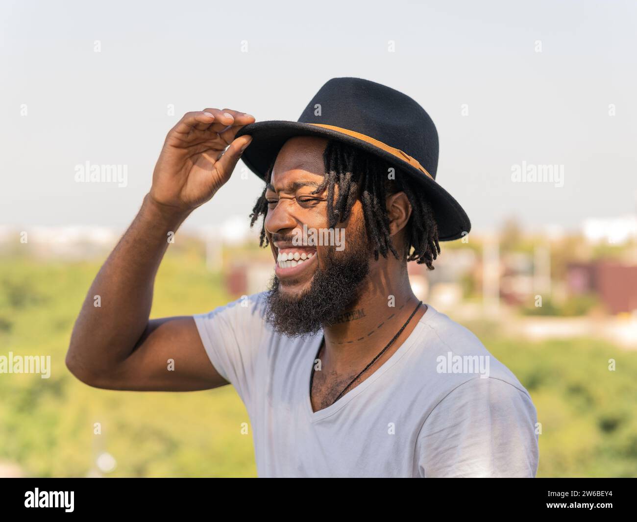 Lächelnder junger bärtiger Afroamerikaner in weißem T-Shirt, der einen schwarzen Hut anpasst und glücklich über das Stadtbild lacht Stockfoto