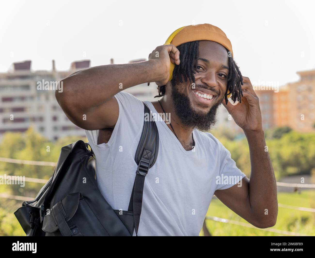 Lächelnder junger afroamerikanischer männlicher Hipster mit Rucksack, der am Sommertag im Stadtpark steht und Kopfhörer berührt und Musik hört Stockfoto