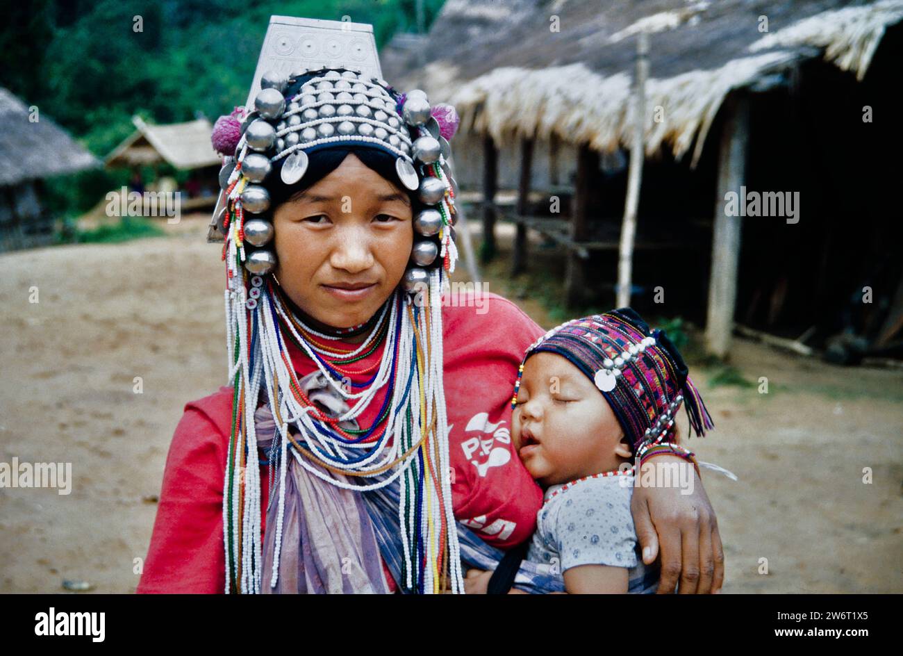 Historisches Archiv Bild einer Akha-Frau in traditioneller Erwachsenenkopfhose aus Münzen und Metallkugeln und Einem Jejaw-Perlen-Schärpe mit Einem schlafenden Baby in Einer traditionellen geflochtenen Kappe mit Blick auf die Kamera mit traditionellem Akha-Dorf im Hintergrund, nördlich von Chiang Mai. Thailand 1990 Stockfoto