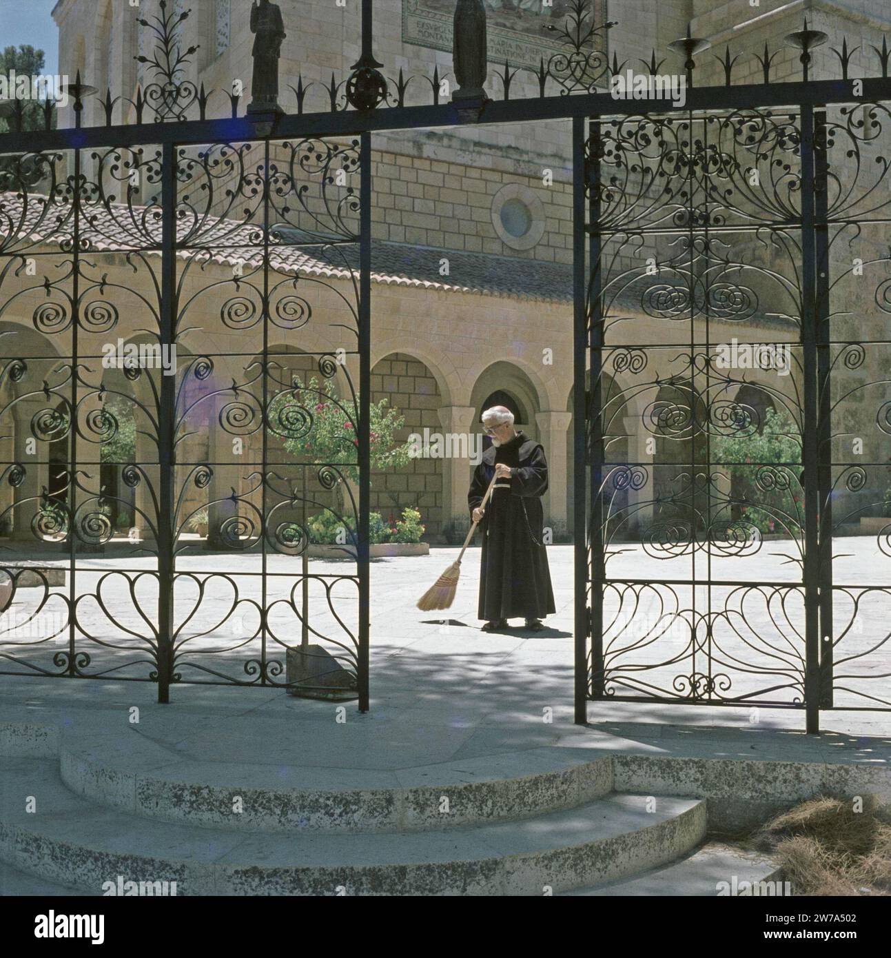 Ein Karam. Kirche der Besichtigung: Ein Franziskanerbruder fegt den Platz in der Nähe der Kirche. Im Vordergrund befindet sich ein schmiedeeisernes Tor ca. 1964 Stockfoto