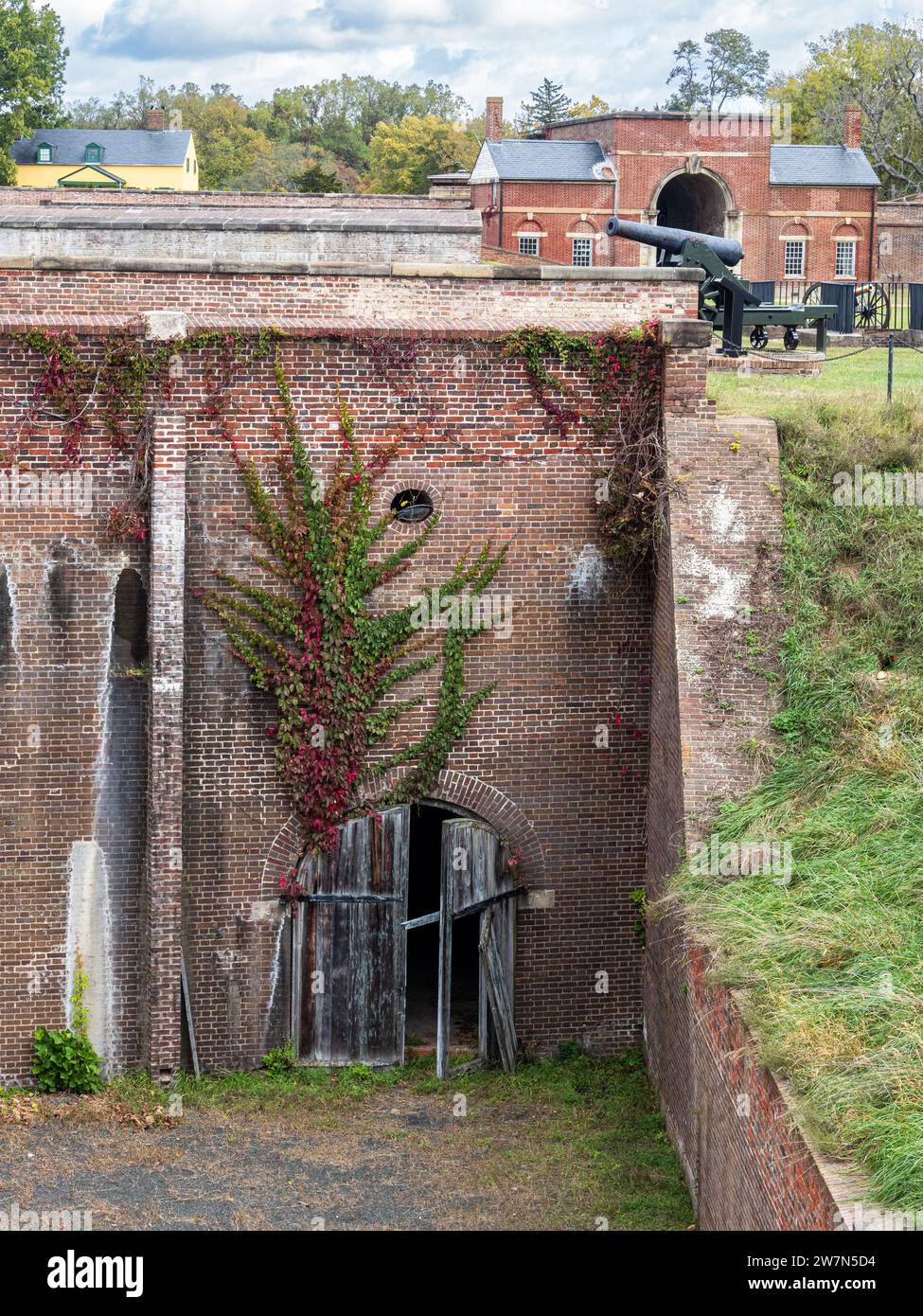 Die verwitterte hölzerne Kellertür eines roten Ziegelbaus, geschmückt mit Kletterstöcken, mit einer Kanone als Hintergrund in Fort Washington, Virginia. Stockfoto