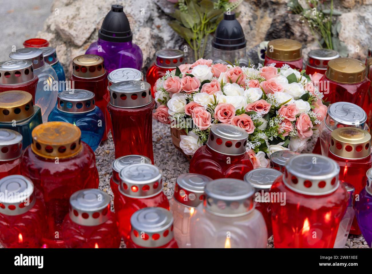 Verschiedene Grabkerzen (Gedenkkerzen) in verschiedenen Farben (blaue, rote, transparente Behälter) auf einem Friedhof; mit Blumen Stockfoto