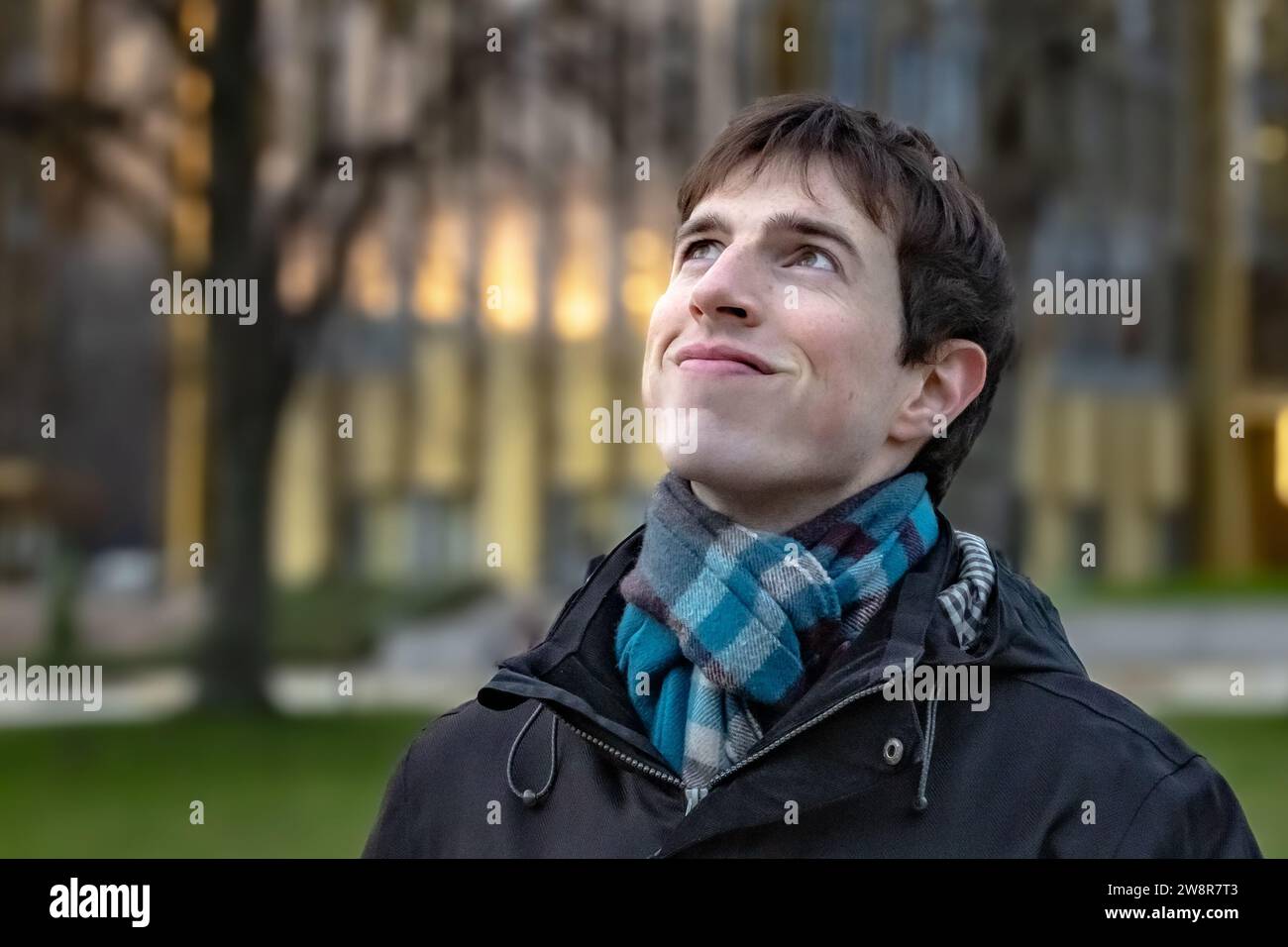Ein junger Mann, der an einem kalten Tag vor einem Universitätsgebäude steht, trägt einen Schal und lächelt, während er nach oben und vielleicht in die Zukunft blickt. Stockfoto