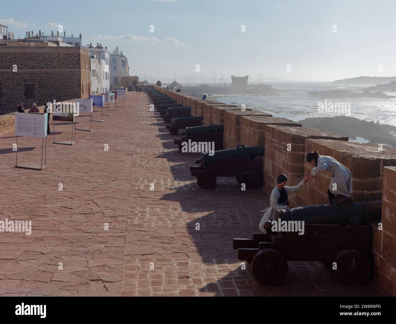 Touristen klettern auf die Kanonenpistolen neben den Stadtmauern mit Blick auf das Meer in Essaouira, auch bekannt als „die windige Stadt“, Marokko. Dezember 2023 Stockfoto