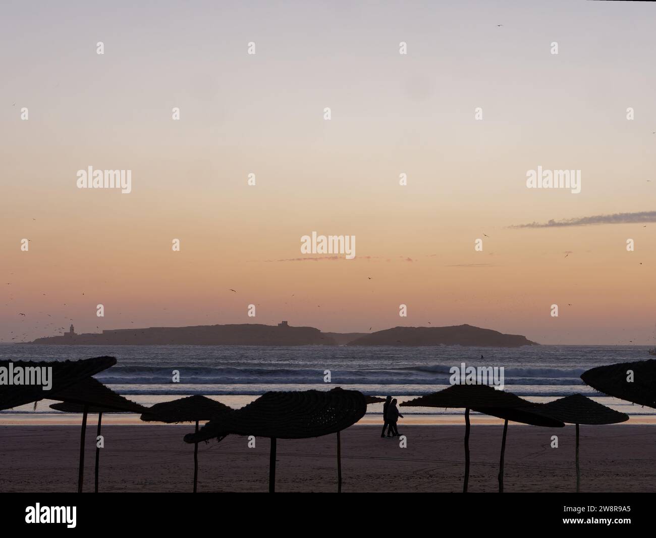 Spazieren Sie zu zweit an einem Sandstrand mit Sonnenschirmen bei Sonnenuntergang mit einer Insel dahinter, in Essaouira 'The Windy City', Marokko. Dezember 2023 Stockfoto