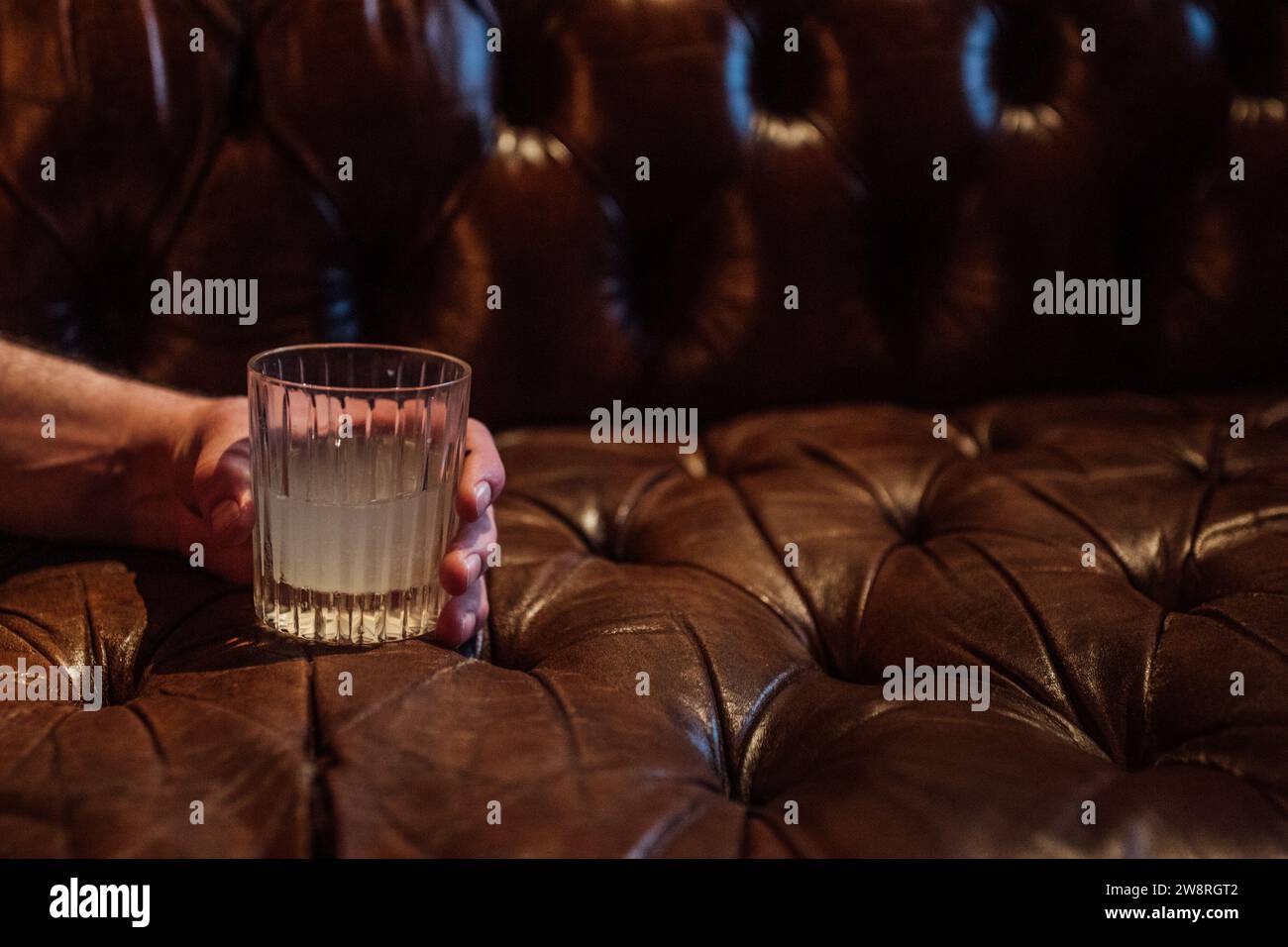 Die Hand des Mannes hält Kristallglas mit einem Cocktail auf der braunen Chesterfield Couch Stockfoto