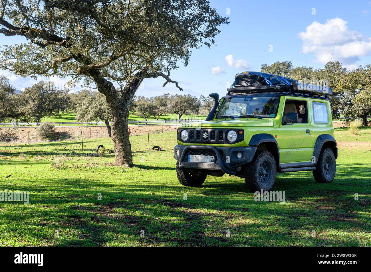 Phosphoreszierender grüner Suzuki Jimny, elektrisch gelb, bereit für Überlandfahrten, beschattet unter einem Baum mitten auf dem Feld, mit einem Dach ausgestattet Stockfoto