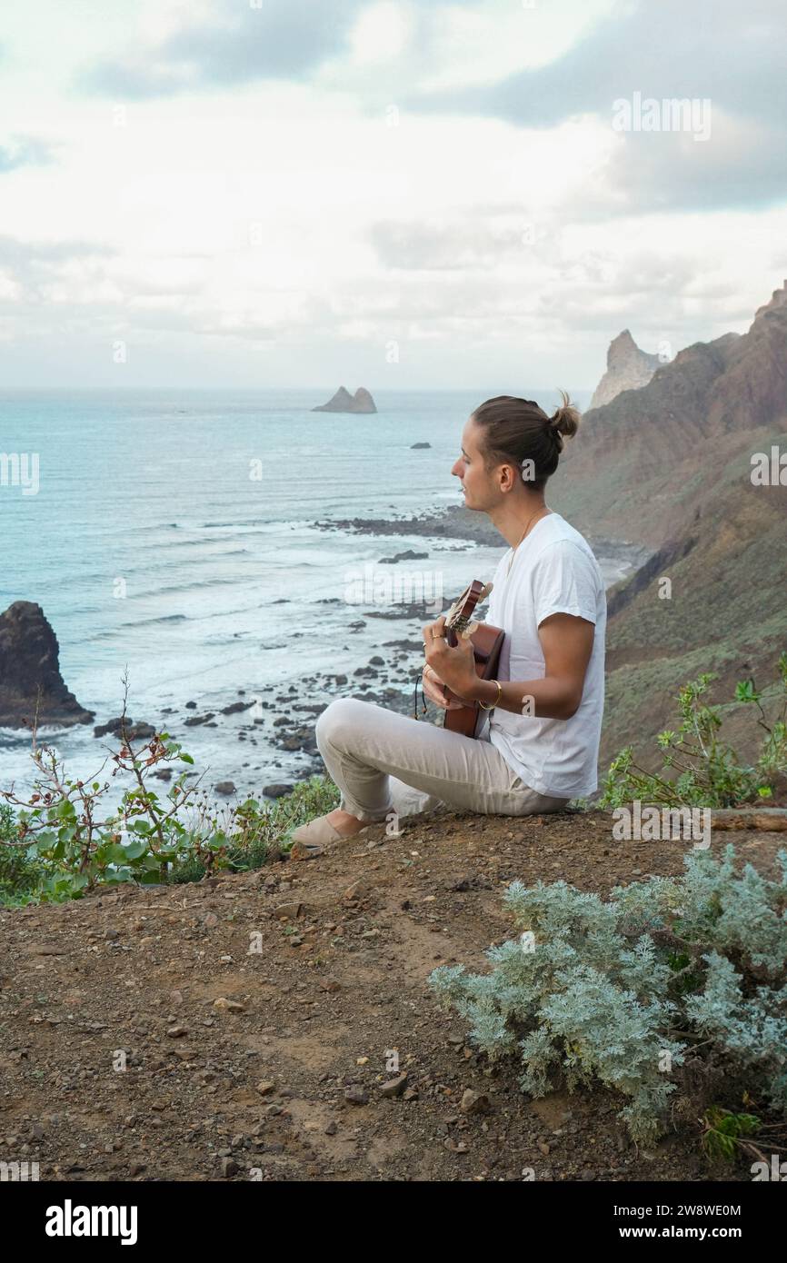Junge, langhaarige Mann in lässiger Kleidung, spielt eine Ukulele an einer Klippe, mit Blick auf das ruhige Meer und zerklüftete Klippen Stockfoto