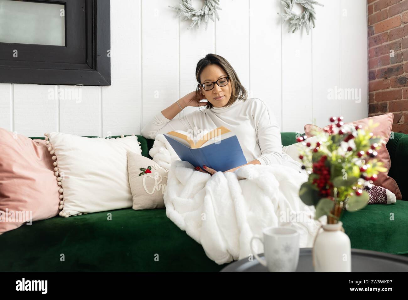 Frau, die auf der Couch sitzt und ein Buch liest, mit Lesebrille Stockfoto