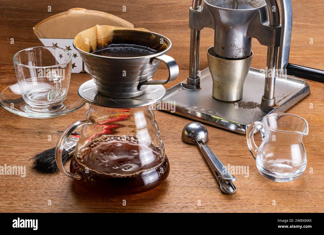 Hochwinkelansicht der Ausrüstung für tropfenden Kaffee, Kaffeemühle, Papierfilter, Filterhalter, Glasbehälter, Löffel und Bürste auf Holztisch. Stockfoto