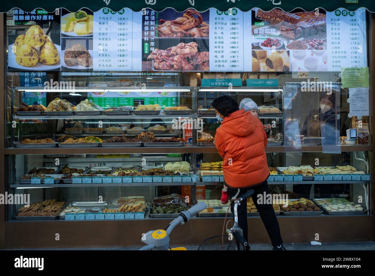 Eine Frau (möglicherweise eine Großmutter) schaut vor einem Lebensmittelgeschäft mit ihrem Kind in den Armen in Peking, China. 22-Dez-2023 Stockfoto