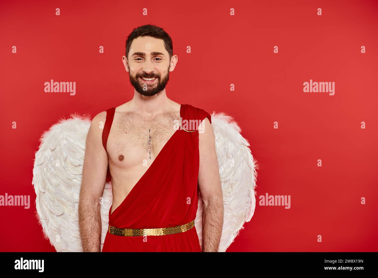 Positiver bärtiger Mann, als Amulett gekleidet, mit Flügeln, die in die Kamera lächeln, am roten Valentinstag Stockfoto