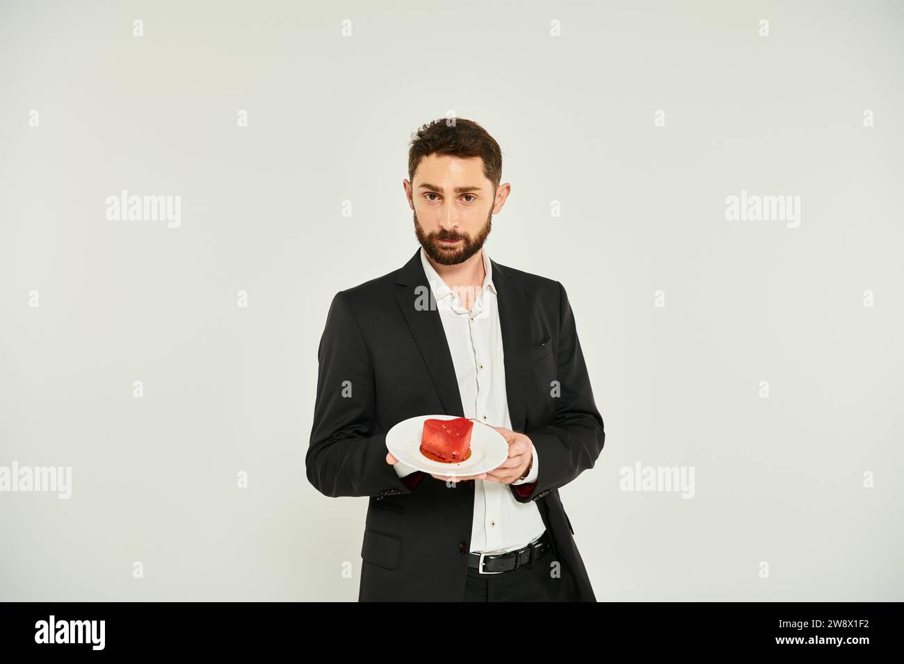 Eleganter bärtiger Mann, der einen Teller mit einem höhlenförmigen Kuchen auf grauem, köstlichem Valentinstag-Leckerbissen zeigt Stockfoto