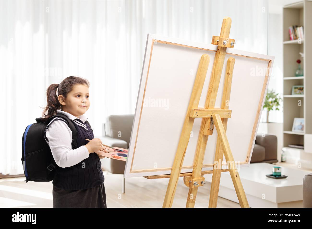 Das Schulmädchen hält eine Farbpalette und einen Pinsel und steht vor einer Leinwand in einem Raum Stockfoto
