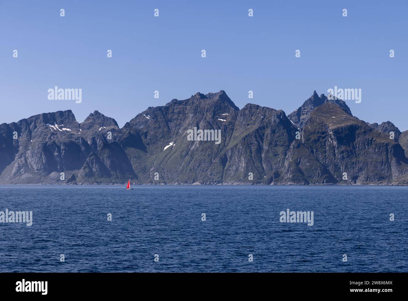 Ein malerischer Blick auf die Nordsee und einen Bergkamm, der über ruhigem Wasser thront, mit einem Segelboot mit rotem Segel nahe der Insel Lofoten, unter einem bl Stockfoto