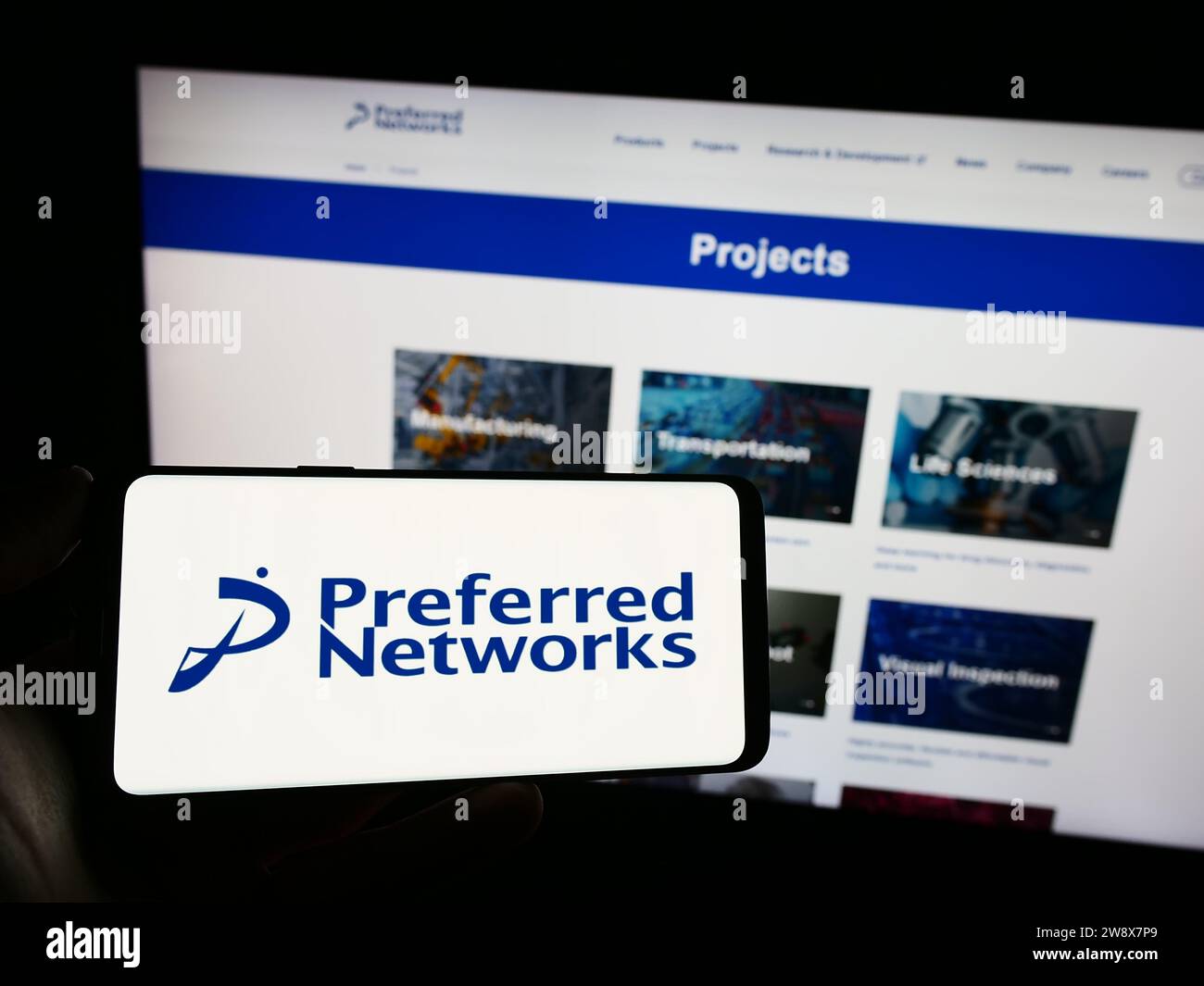 Person, die ein Mobiltelefon mit dem Logo des Unternehmens Preferred Networks Inc. Für künstliche Intelligenz vor der Webseite hält. Schwerpunkt auf dem Display des Telefons. Stockfoto