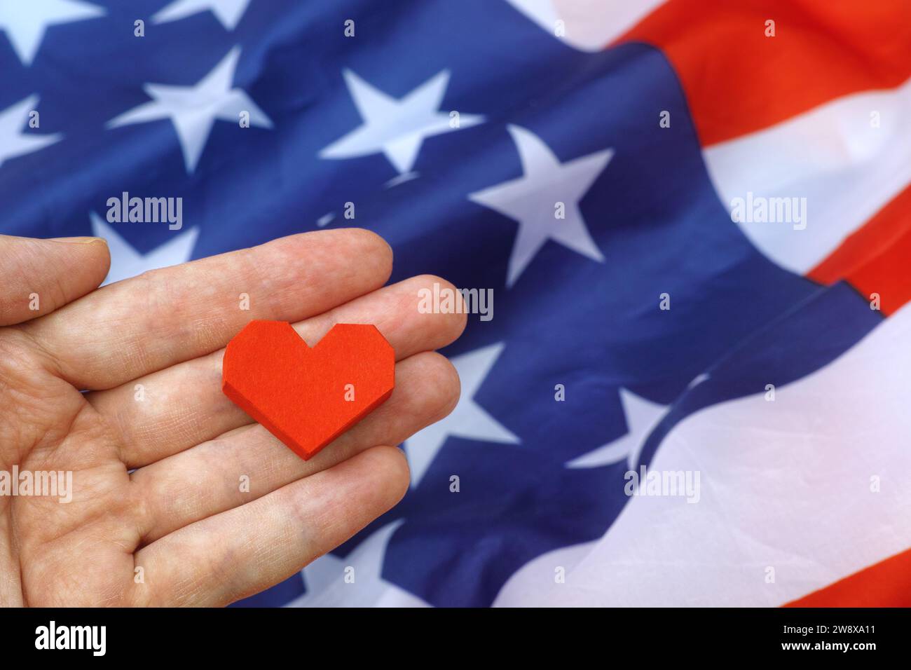 Eine Person, die ein rotes Origami-Herz über der Flagge der USA hält. Nahaufnahme. Stockfoto