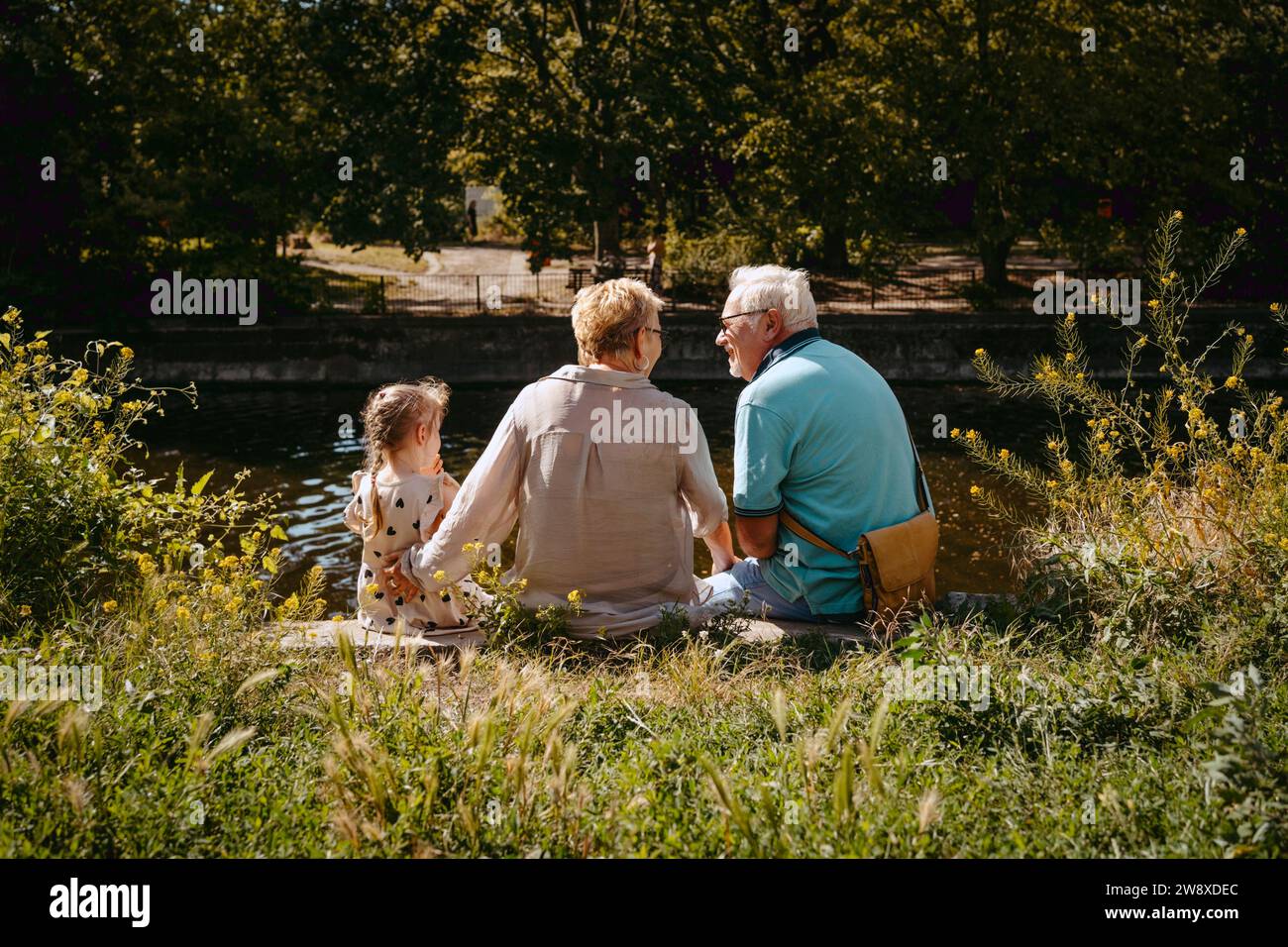 Rückansicht der Großeltern, die mit Enkelin am Teich im Park sitzen Stockfoto