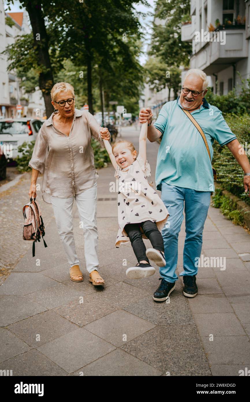 Verspielte Großeltern genießen es mit Enkelin, während sie auf einem Fußweg an der Straße spazieren gehen Stockfoto
