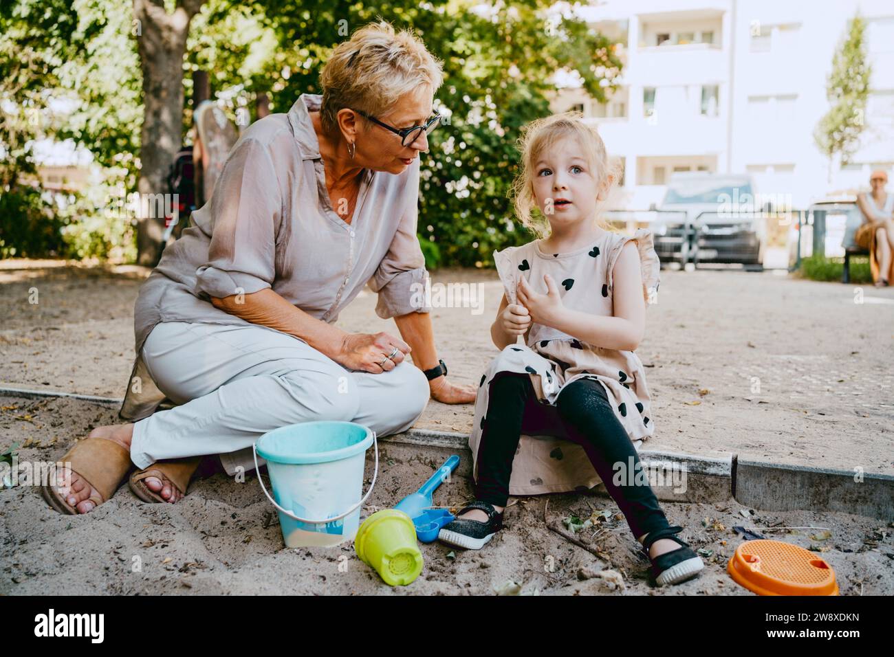 Großmutter spricht mit Enkelin, die im Sandkasten sitzt, während sie im Park spielt Stockfoto