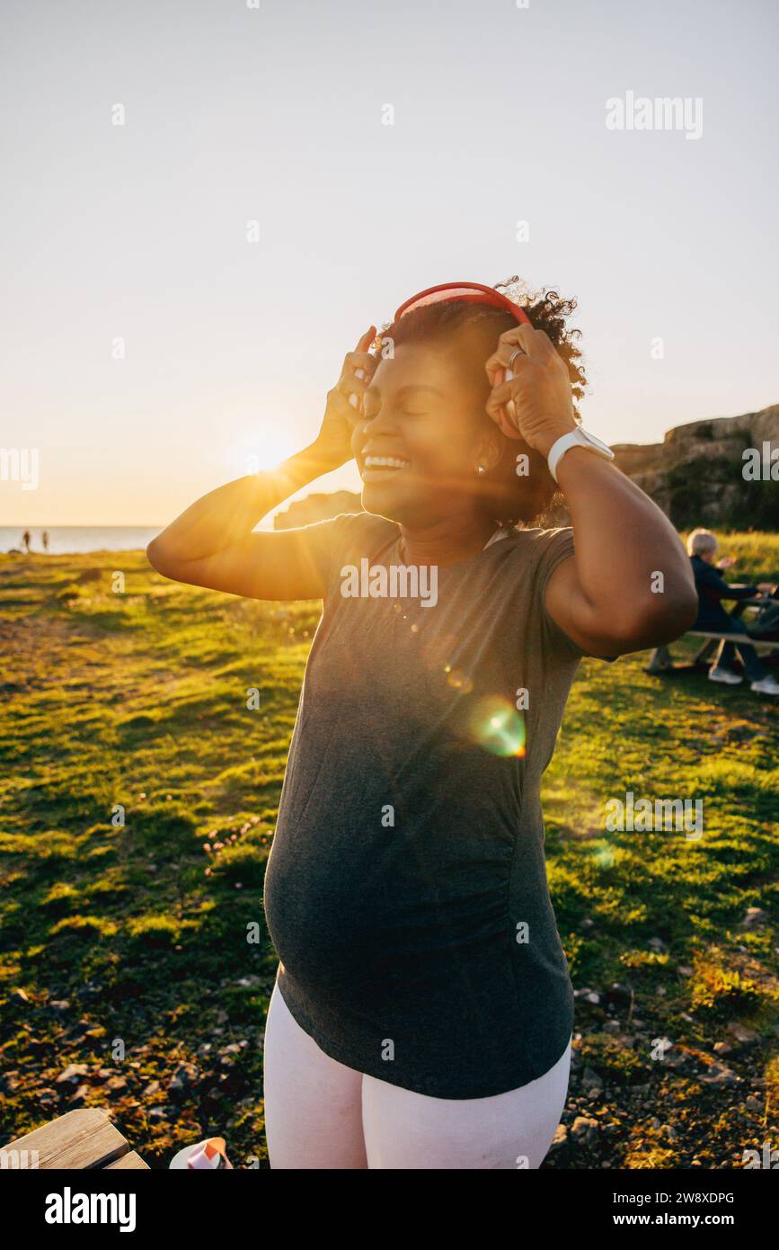 Glückliche schwangere Frau, die bei Sonnenuntergang kabellose Kopfhörer trägt Stockfoto