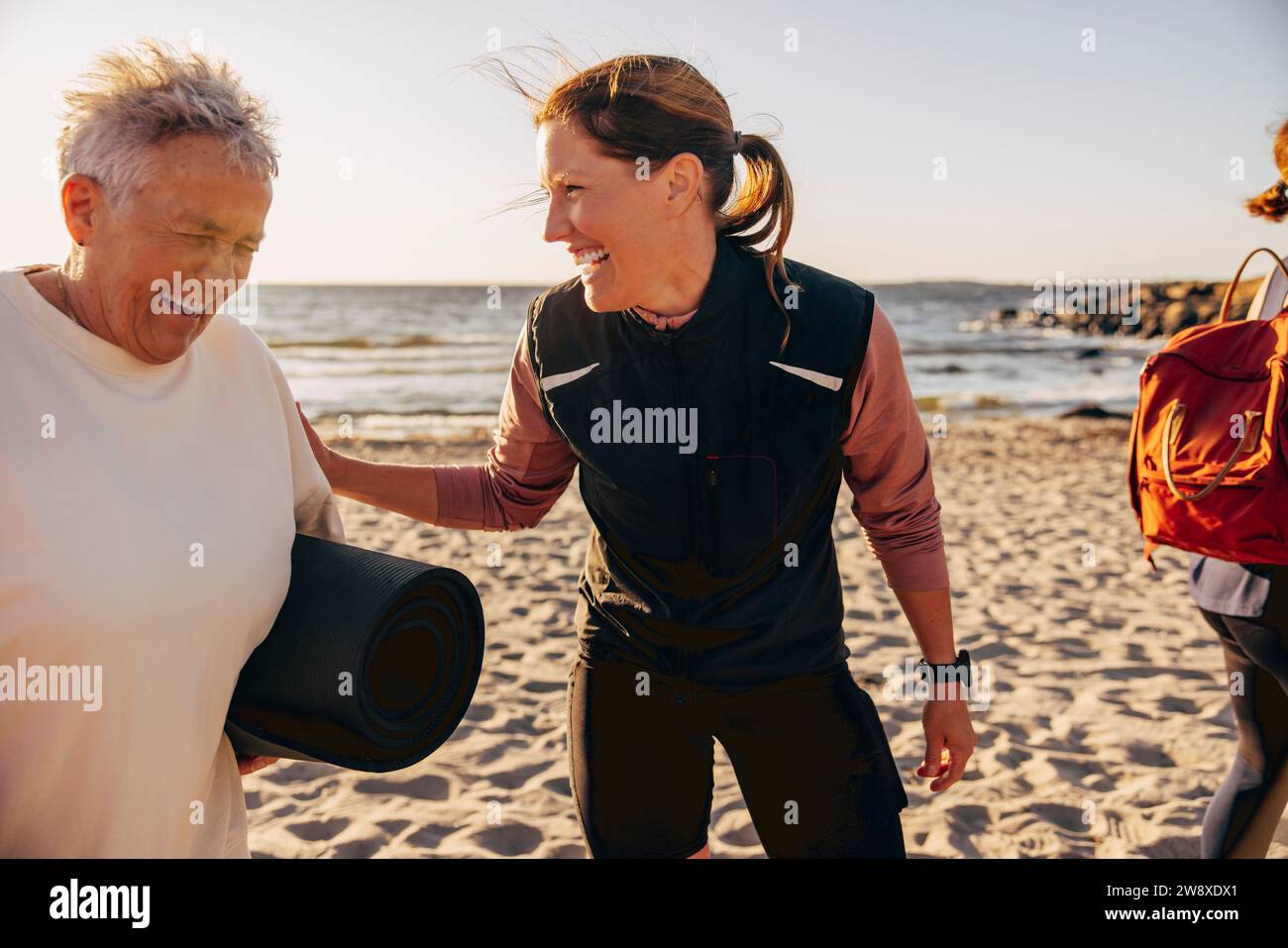 Glückliche Trainerin lacht mit einer älteren Frau, die am Strand eine Trainingsmatte hält Stockfoto