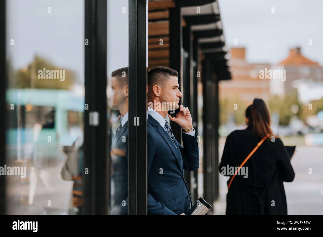 Geschäftsmann, der mit dem Smartphone spricht, während er sich auf der Straße an eine Glastür lehnt Stockfoto