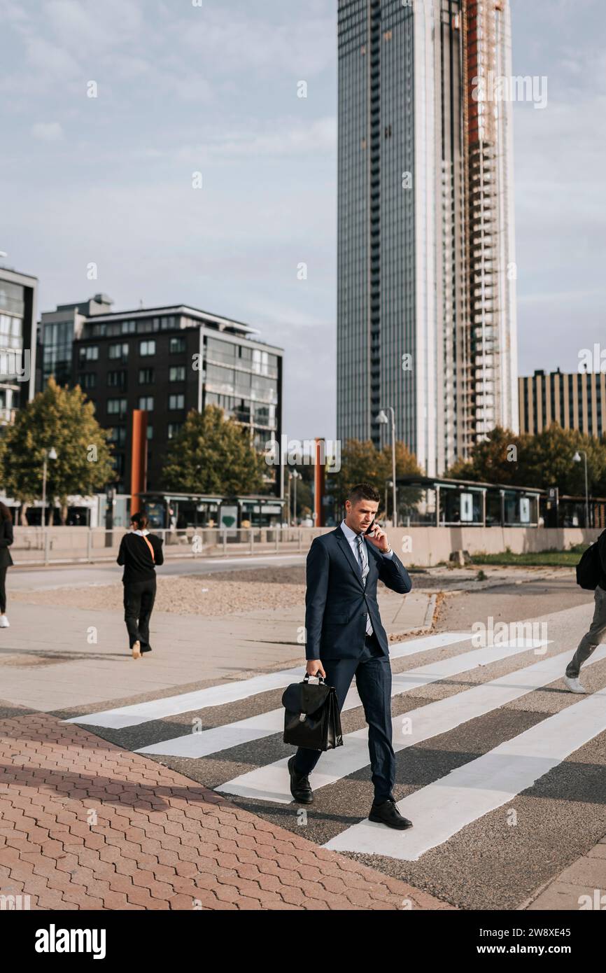 Geschäftsmann, der über das Smartphone spricht, während er auf der Straße in der Stadt auf einem Zebraübergang läuft Stockfoto