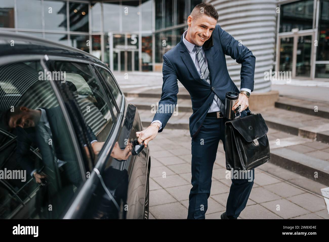 Geschäftsmann, der mit dem Smartphone spricht, während er die Autotür in der Nähe des Bürogebäudes öffnet Stockfoto