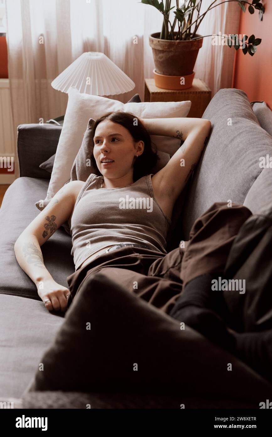 Junge Frau mit erhobener Hand, die wegschaut, während sie sich auf dem Sofa in der Wohnung liegend Stockfoto