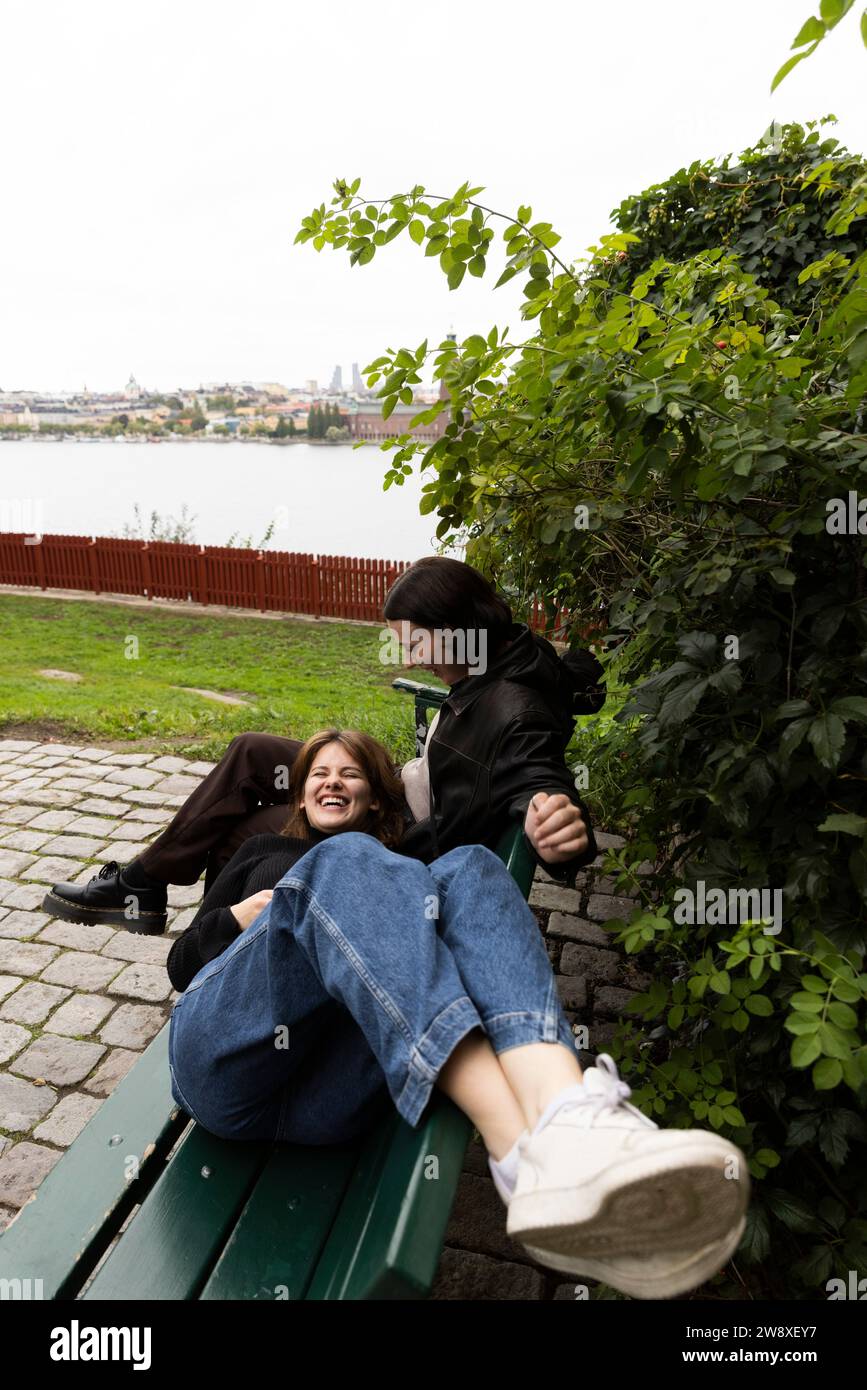 Glückliche Frau, die auf dem Schoß einer Freundin liegt, die auf einer Bank im Park sitzt Stockfoto