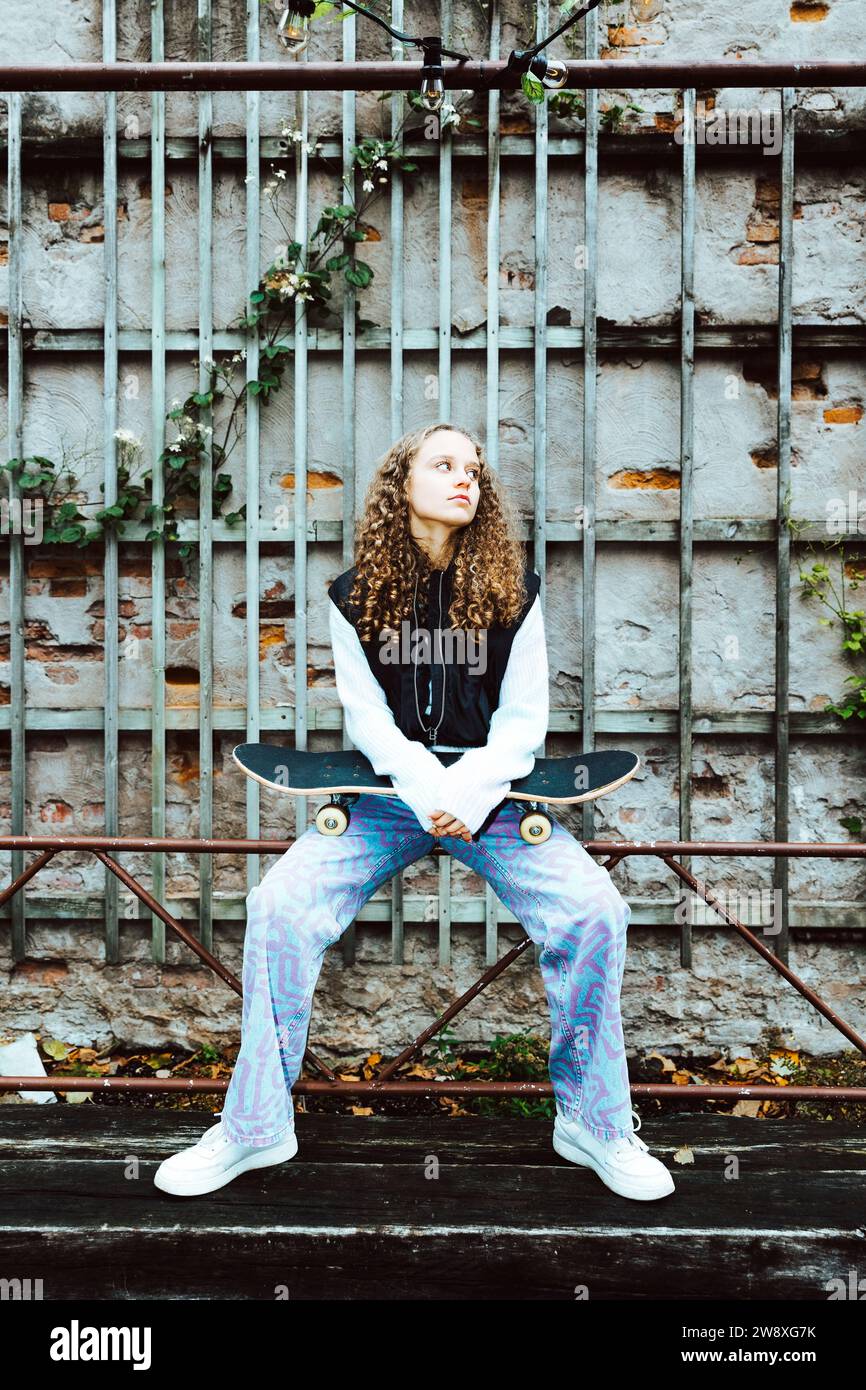 Besinnliches Mädchen mit Skateboard, das auf einem Geländer gegen die Wand sitzt Stockfoto