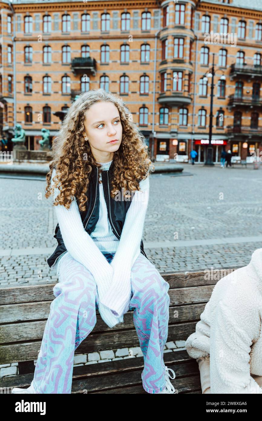 Ein besinnliches Mädchen, das wegschaut, während er am Wochenende in der Stadt auf der Bank sitzt Stockfoto