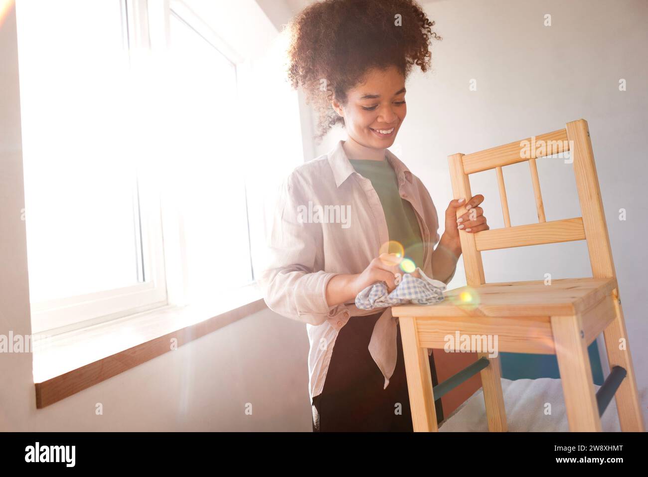 Ein wunderschönes, lächelndes Mischlingsmädchen wischt einen Holzstuhl mit einem Lappen ab. Eine attraktive junge Afrikanerin bereitet Möbel zum Malen vor. Renovierung Stockfoto