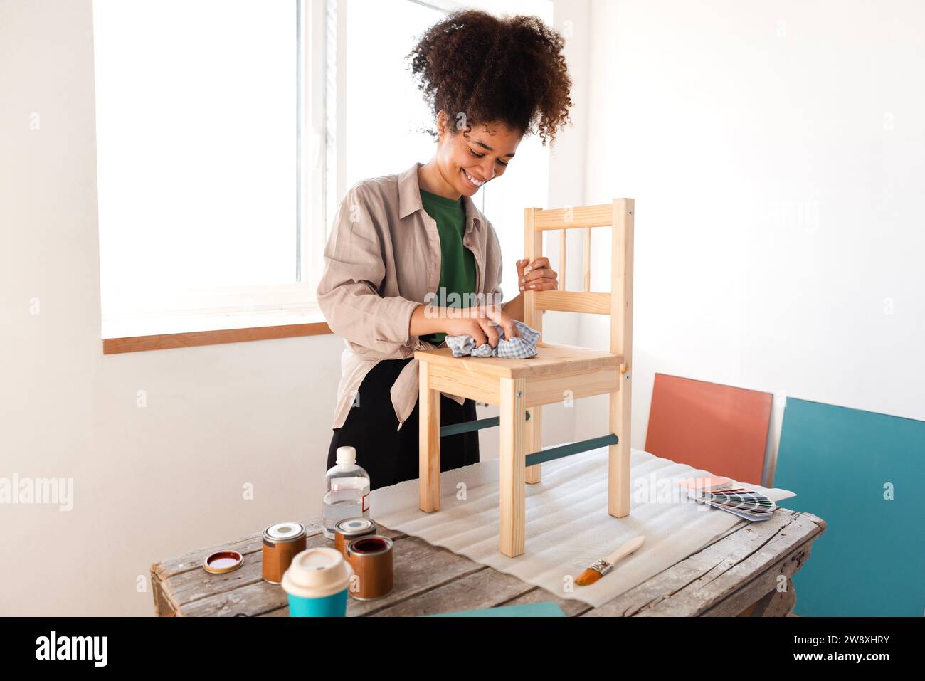 Ein wunderschönes, lächelndes afro-amerikanisches Mädchen wischt einen Holzstuhl mit einem Lappen ab. Eine attraktive junge Afrikanerin bereitet Möbel zum Malen vor. Renovat Stockfoto