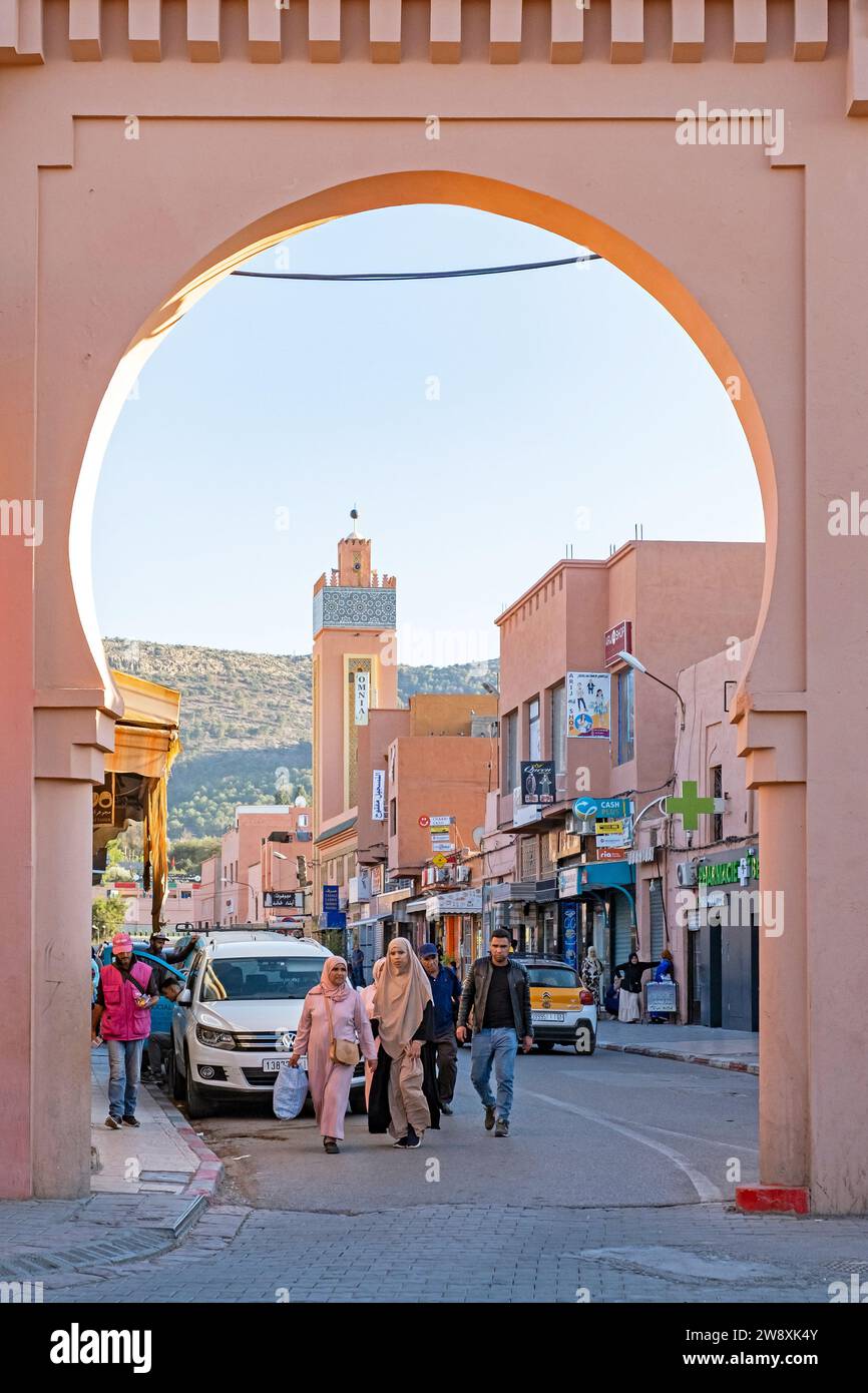 Minarett und Marokkaner, die unter dem pinkfarbenen Stadttor in der Stadt Beni Mellal, Region Béni Mellal-Khénifra, Marokko spazieren Stockfoto
