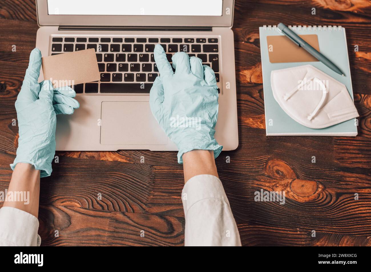 Bild der Hände in medizinischen Handschuhen auf dem Hintergrund eines Laptops. Die Frau hält eine Plastikkarte und gibt die Nummer auf der Website ein. Online Stockfoto