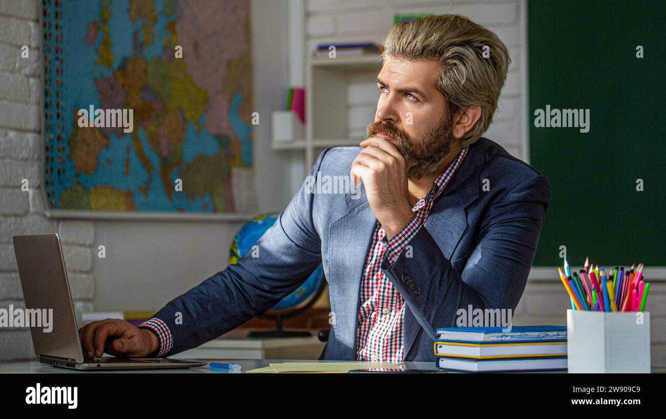Ein Lehrer mit Laptop in der Klasse sitzt am Tisch, an der Tafel im Klassenzimmer. Lehrer nachdenklich im Klassenzimmer der Grundschule Stockfoto