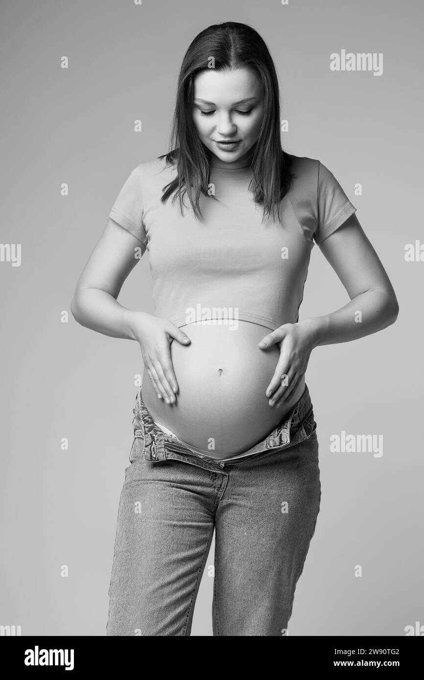 Schwarz-weißes Porträt einer schwangeren Frau in T-Shirt und Jeans auf grauem Hintergrund. Frau, die auf den schwangeren Bauch blickt. Bauch freigelegt. 6. Monat Preg Stockfoto