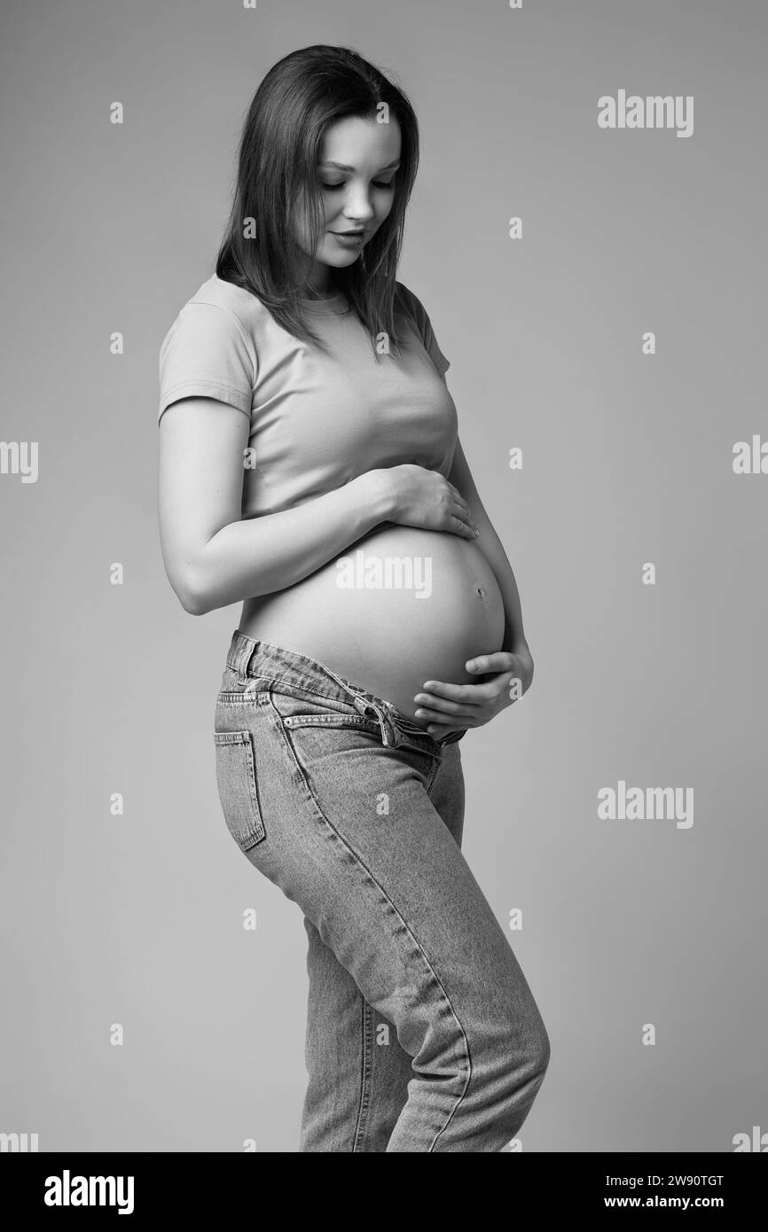 Schwarz-weißes Porträt der jungen hübschen schwangeren Frau in T-Shirt und Jeans auf grauem Hintergrund. Frau, die auf den schwangeren Bauch blickt. Bauch freigelegt. 6 Stockfoto