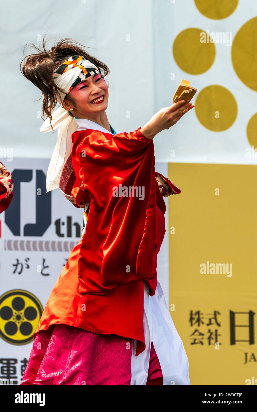 Die junge, lächelnde, glückliche japanische yosakoi-Tänzerin tanzt auf der Bühne in einer hellroten Yukata-Tunika mit langen Ärmeln. Hält eine Naruko-Holzklatsche aus. Stockfoto