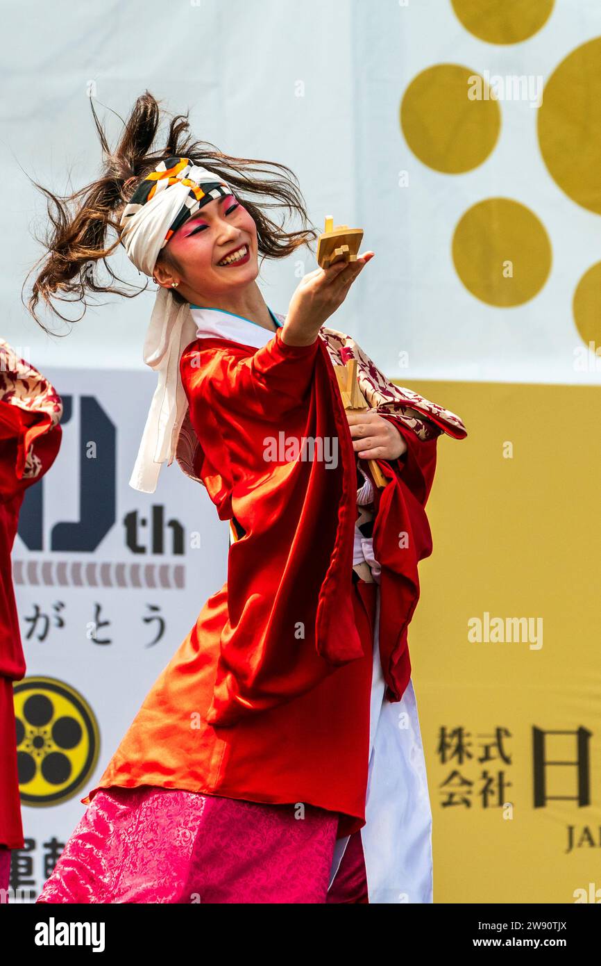 Die junge, lächelnde, glückliche japanische yosakoi-Tänzerin tanzt auf der Bühne in einer hellroten Yukata-Tunika mit langen Ärmeln. Hält eine Naruko-Holzklatsche aus. Stockfoto