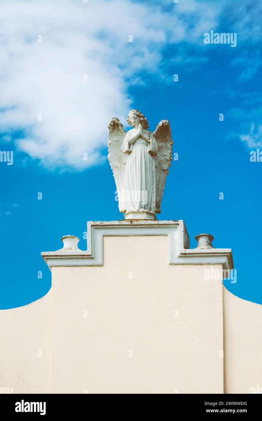 Weibliche Skulptur eines betenden Engels auf einer weißen Wand mit Kopierraum, mit blauem Himmel mit Wolken im Hintergrund Stockfoto