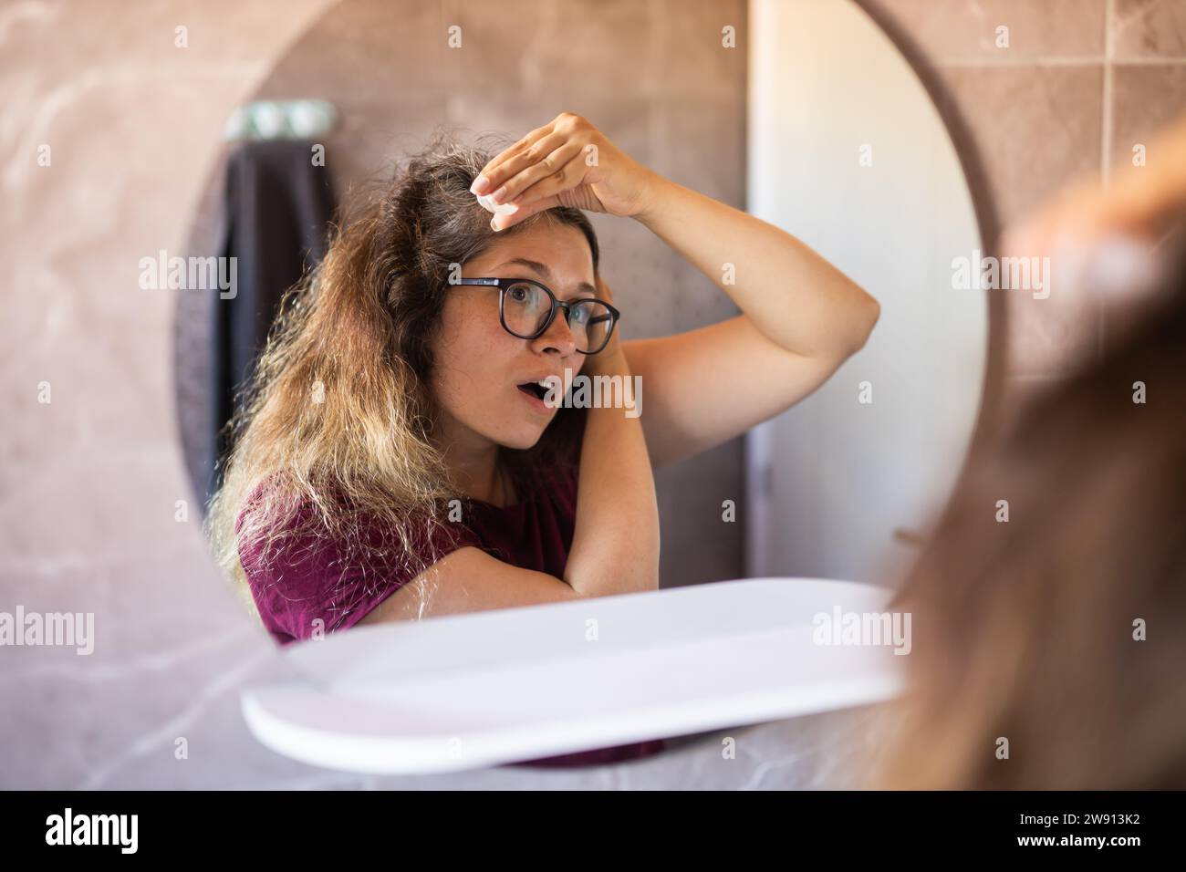 Grauhaarige überraschte kaukasische Frau mittleren Alters, die den grauen Haarkopf in Spiegelreflektion betrachtet. Frühes graues Haarkonzept Stockfoto