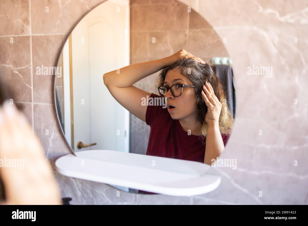 Grauhaarige überraschte kaukasische Frau mittleren Alters, die den grauen Haarkopf in Spiegelreflektion betrachtet. Frühes graues Haarkonzept Stockfoto