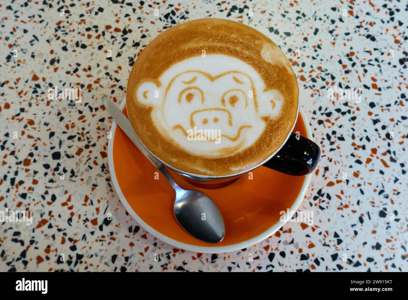 Eine Tasse flacher weißer Kaffee mit einem Affengesicht im Schaum darüber in einer Tasse mit Teelöffel und Untertasse. Stockfoto
