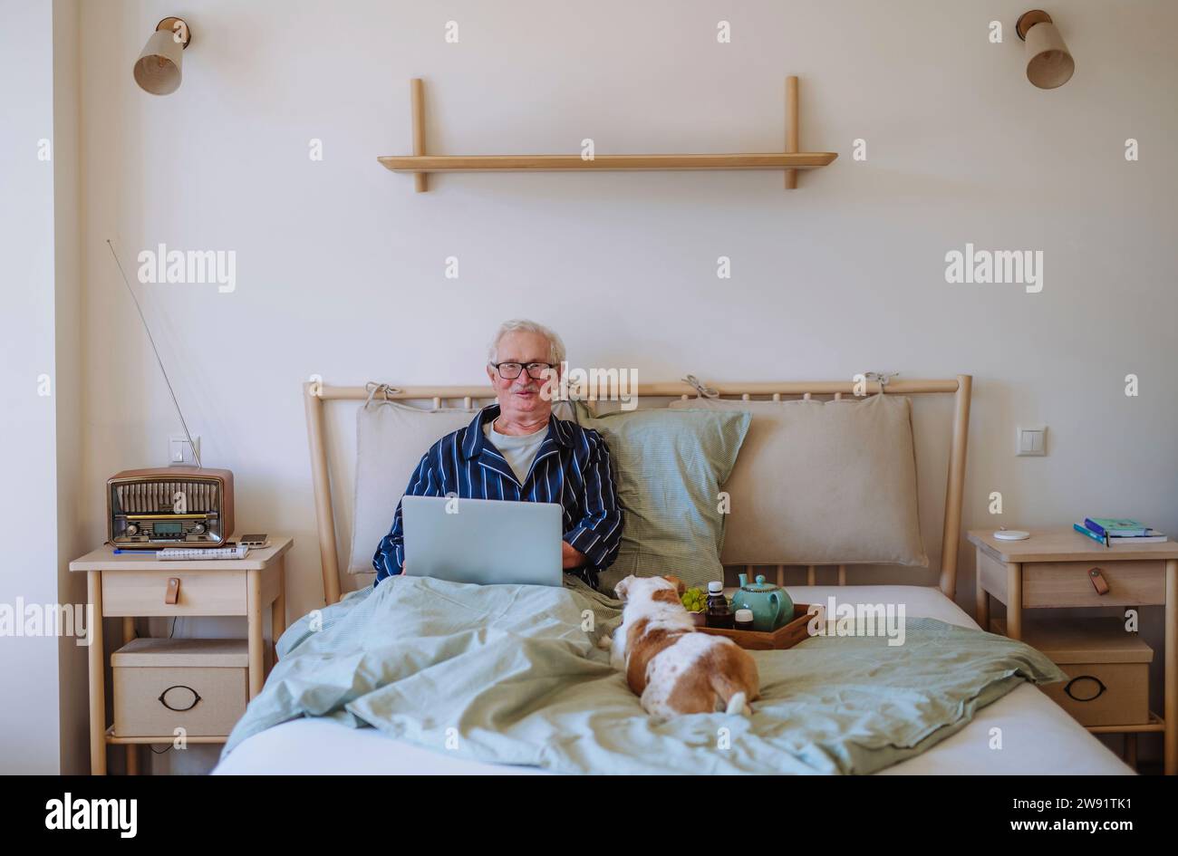 Ein lächelnder Seniorenmann sitzt mit Laptop und Hund zu Hause auf dem Bett Stockfoto