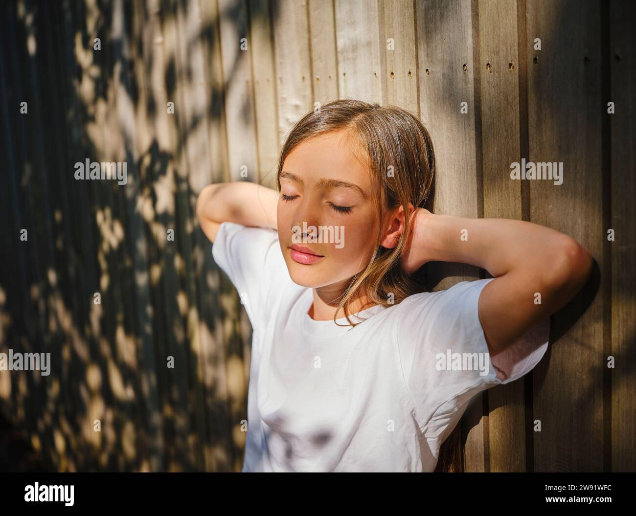 Mädchen mit geschlossenen Augen, lehnt sich an eine Holzwand und genießt Sonnenlicht Stockfoto