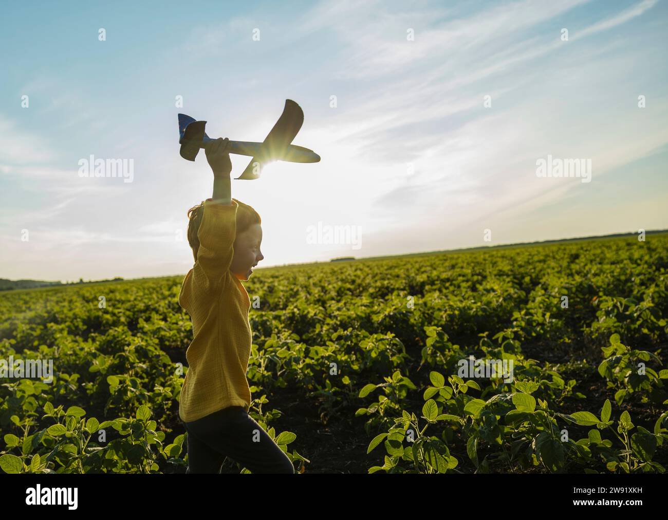 Fröhlicher Junge, der mit einem Spielzeugflugzeug auf dem landwirtschaftlichen Feld unter dem Himmel läuft Stockfoto