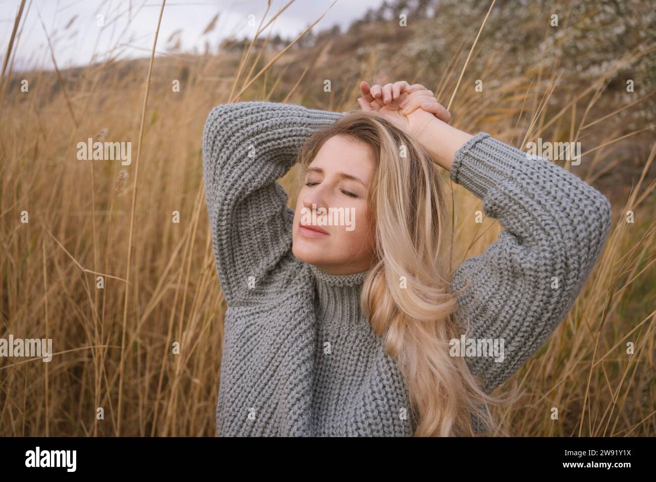 Blonde Frau, die einen grauen Pullover in der Nähe von trockenem, hohem Gras trägt Stockfoto